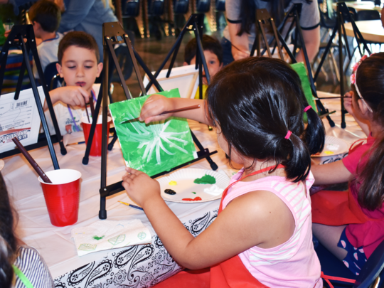 A group of young campers experiment in a painting class. In the foreground is back of a young girl who is focused on a while flower on a green field.