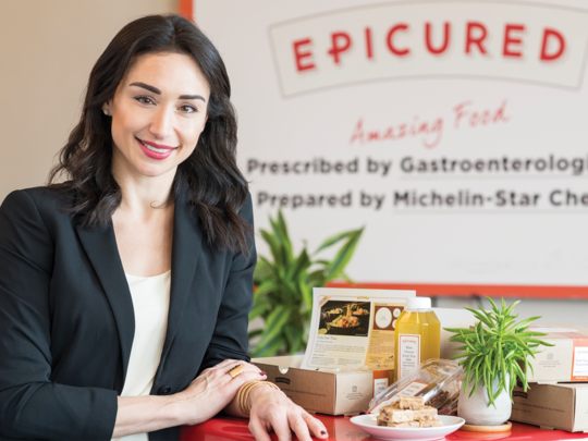An Armenian woman in a black blazer, smiling and resting her arm on a table with an assortment of gourmet food on it. The woman is smiling and the logo of her company "Epicured" is in the background.